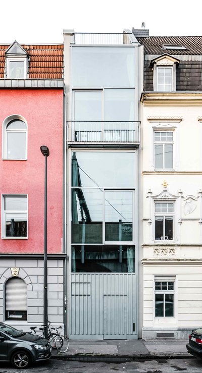 Schmales modernes Gebäude mit Glasfassade zwischen einem rosa und einem weißen Altbau in einer städtischen Straßenszene mit parkenden Autos.