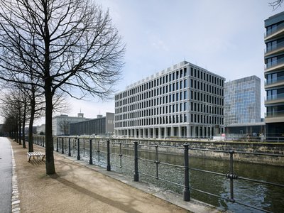 Modernes Bürogebäude aus Beton am Ufer eines Kanals in Berlin mit Bäumen und einem Weg im Vordergrund.
