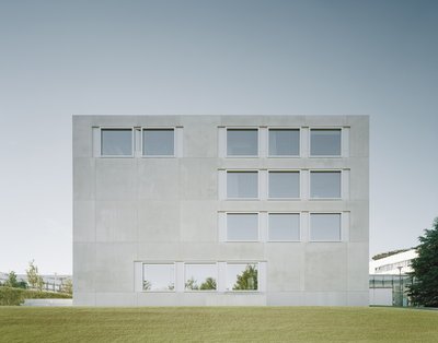 Ein modernes, kubisches Seminarhaus aus hellem Beton mit regelmäßigen Fensterreihen steht auf einer Wiese unter klarem Himmel.