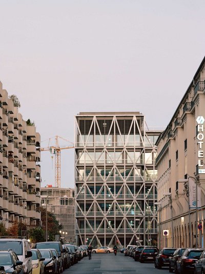 Straßenszene mit modernem Glasbau im Zentrum, flankiert von geparkten Autos und Wohnhäusern unter einem bedeckten Himmel.