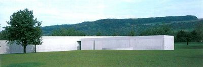 Langes, weißes Betonbauwerk auf einer grünen Wiese mit Baum links im Vordergrund und bewaldetem Hügel im Hintergrund unter hellem Himmel.
